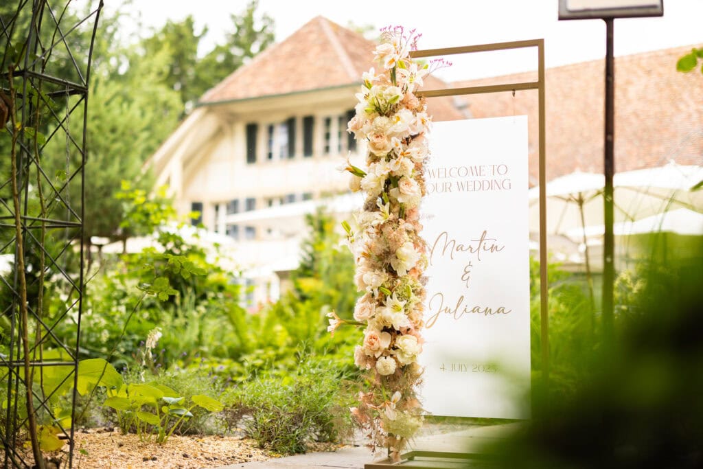 Elegant wedding welcome sign with floral arrangement in lush garden setting.